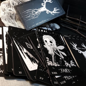 May include: A deck of black tarot cards with white illustrations. The cards feature a variety of images, including a fairy, a skull, and a deer. The cards are numbered with Roman numerals and have the word "Fairy" written on one of the cards.