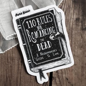 Puede incluir: Una pegatina en blanco y negro con un diseño de libro vintage. El libro se titula "110 reglas para enamorar a los muertos: Una guía del nigromante para el amor."