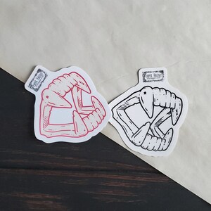 May include: Two sticker designs of a set of fangs, one in red and one in black and white. The stickers are on a white background.