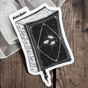 Puede incluir: Pegatina en blanco y negro de un libro cerrado con una forma de diamante negro en la portada. El diamante tiene un patrón de estrellas y tres ojos en su interior. El libro tiene un lomo con una serie de protuberancias y el texto "PAPER HAUNT" en la portada.