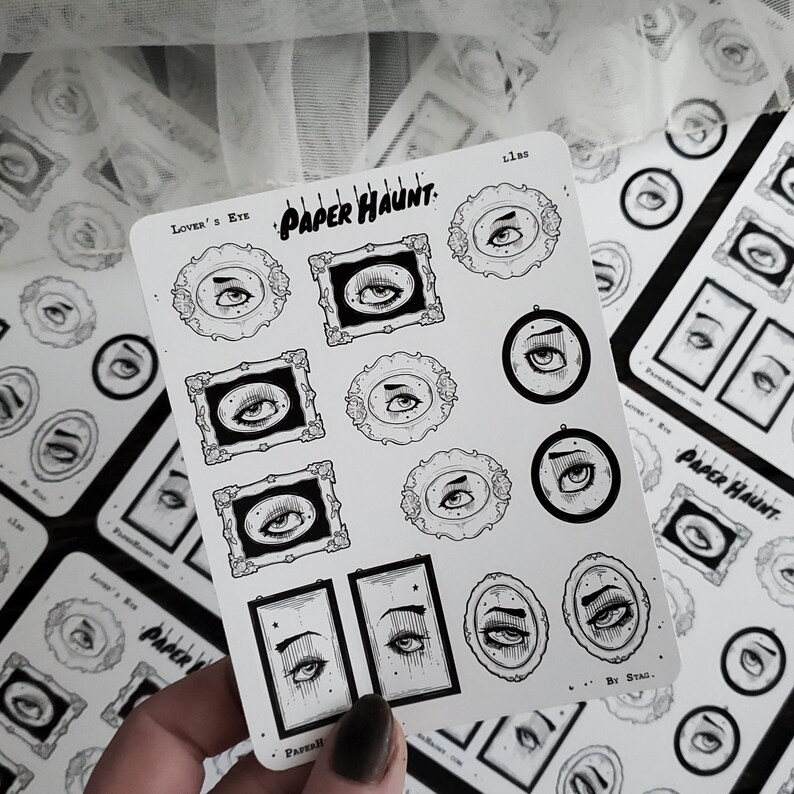 May include: A sheet of black and white stickers featuring various eye designs. The stickers are arranged in a grid pattern and include a variety of styles, including eyes with eyelashes, eyes with stars, and eyes with frames. The stickers are labeled "Paper Haunt" and "Lover's Eye".