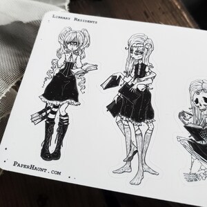 May include: Black and white illustration sticker sheet featuring three characters. The characters are dressed in gothic clothing and holding books. The text "Library Residents" and "PaperHaunt.com" are printed on the sheet.