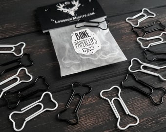 May include: A bag of white bone-shaped paperclips with a sticker that says "Bone Paperclips" and a skull logo. There are also black bone-shaped paperclips scattered on a dark wood surface.