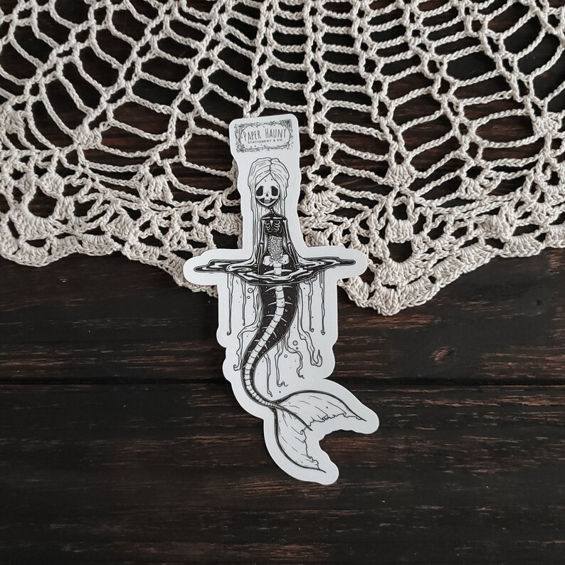 Spooky cute gothic Jelly bones mermaid skeleton emerging from water, a sticker Featuring a Mermaid with Translucent skin with visible skeleton and organs displayed on a doily.