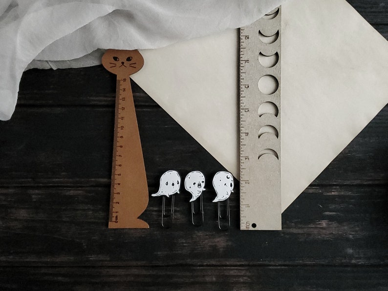 May include: A wooden cat-shaped ruler with a black face and a white background. The ruler is 12 inches long and has markings for each inch. There are three white ghost-shaped paper clips on a dark wooden surface.