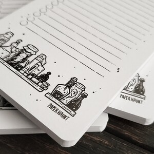 Puede incluir: Un bloc de notas blanco con papel rayado negro y una ilustraci&oacute;n en blanco y negro de botellas de pociones en la parte delantera. El bloc de notas tiene el texto "PAPER HAUNT" impreso en la parte delantera.