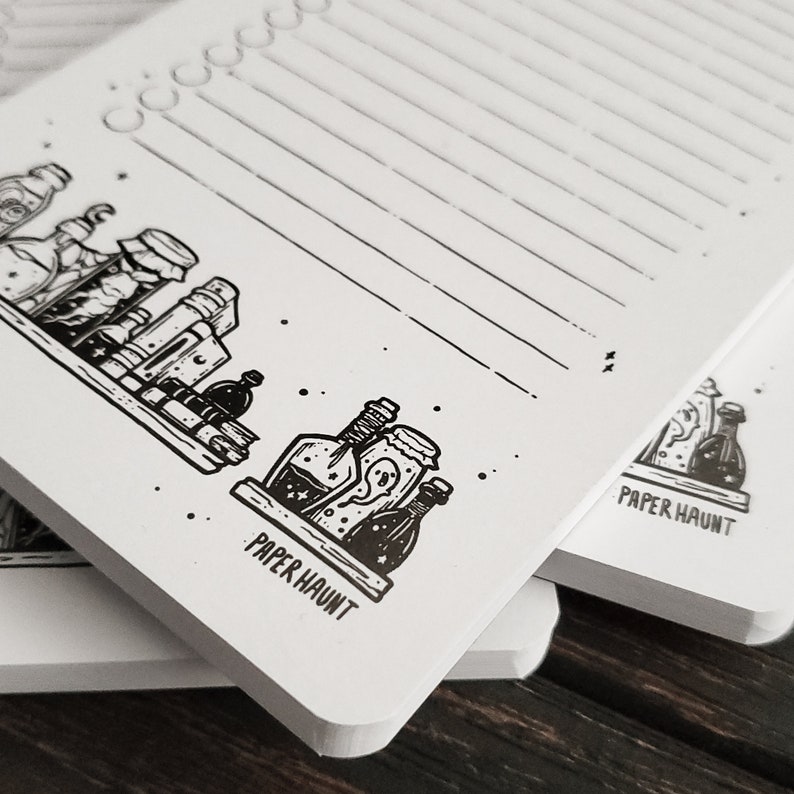 Puede incluir: Un bloc de notas blanco con papel rayado negro y una ilustraci&oacute;n en blanco y negro de botellas de pociones en la parte delantera. El bloc de notas tiene el texto "PAPER HAUNT" impreso en la parte delantera.