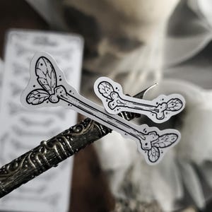May include: Two white stickers with black line art of a bone with leaf-like details. The stickers are on a decorative metal object. A sheet of paper with a pattern is in the background.