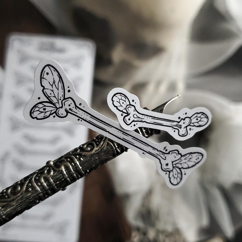 May include: Two white stickers with black line art of a bone with leaf-like details. The stickers are on a decorative metal object. A sheet of paper with a pattern is in the background.