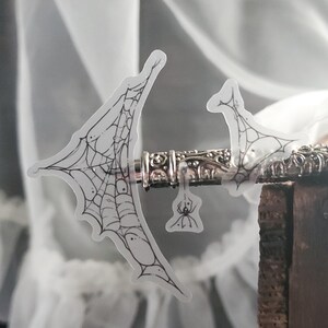 May include: A clear sticker featuring a black and white spiderweb design. The sticker is attached to a silver metal object with intricate details. A small spider is also included on the sticker.