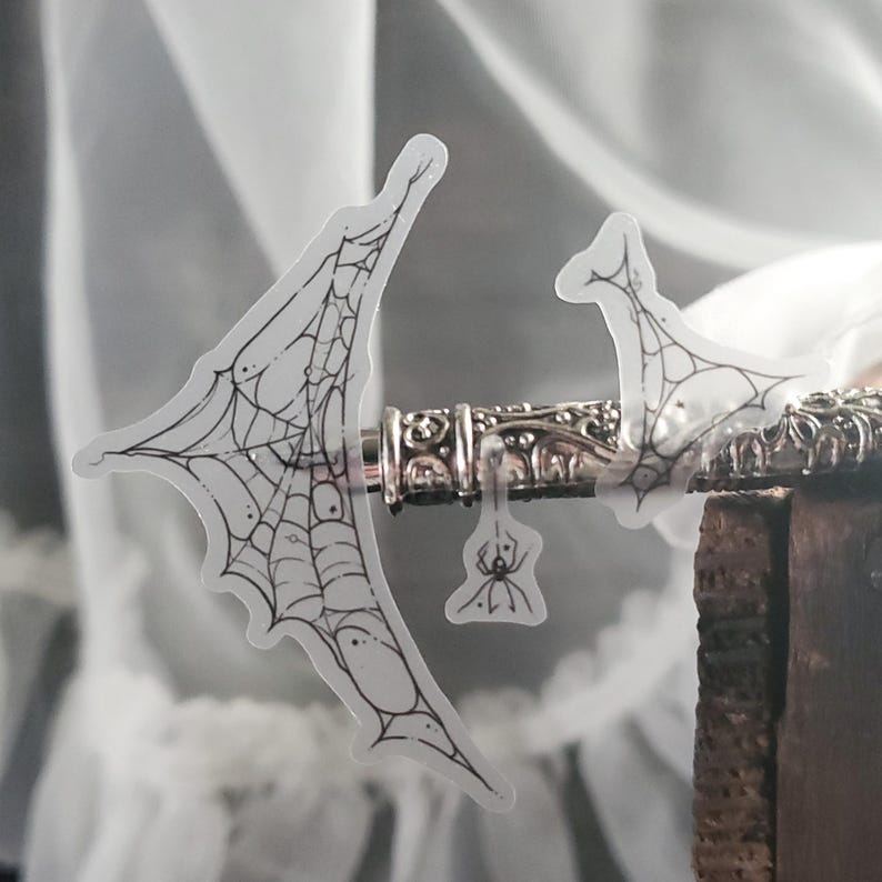 May include: A clear sticker featuring a black and white spiderweb design. The sticker is attached to a silver metal object with intricate details. A small spider is also included on the sticker.