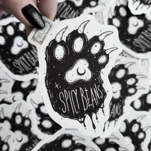 Puede incluir: Pegatina en blanco y negro con un dise&ntilde;o de huella de oso y el texto "Spicy Beans". La pegatina tiene una est&eacute;tica grunge con detalles de luna creciente y estrellas. La pegatina est&aacute; sujeta por una mano con esmalte de u&ntilde;as negro.