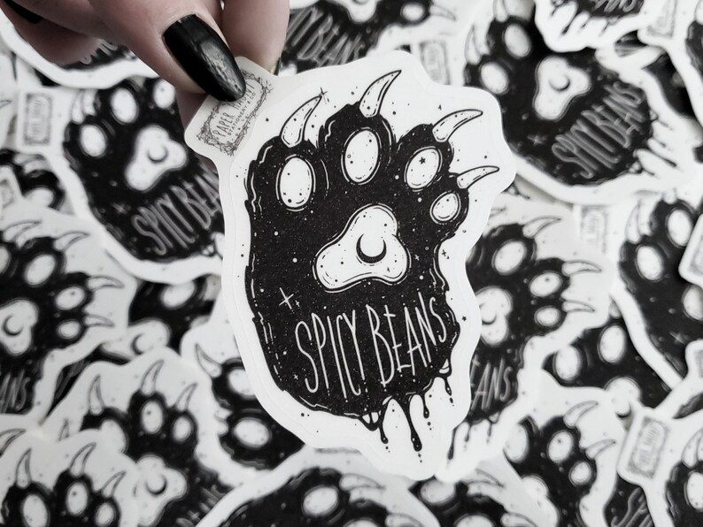 Puede incluir: Pegatina en blanco y negro con un dise&ntilde;o de huella de oso y el texto "Spicy Beans". La pegatina tiene una est&eacute;tica grunge con detalles de luna creciente y estrellas. La pegatina est&aacute; sujeta por una mano con esmalte de u&ntilde;as negro.