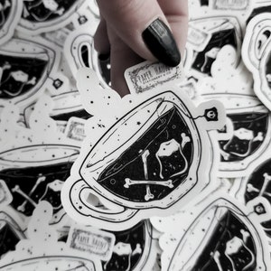Op de afbeelding: Een zwart-witte illustratie van een theekopje met een theezakje en botten erin. Het theekopje is omringd door andere stickers met hetzelfde ontwerp. De tekst "PAPER HAUNT" is zichtbaar op een van de stickers.