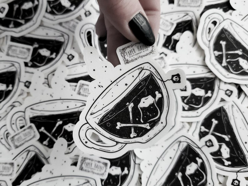 Op de afbeelding: Een zwart-witte illustratie van een theekopje met een theezakje en botten erin. Het theekopje is omringd door andere stickers met hetzelfde ontwerp. De tekst "PAPER HAUNT" is zichtbaar op een van de stickers.