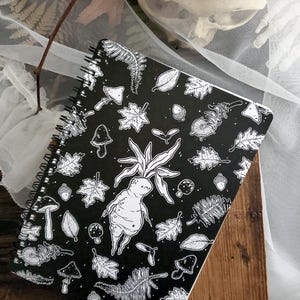 May include: A black spiral notebook featuring a white and black illustration of a woodland scene. The illustration includes mushrooms, leaves, acorns, and a creature with a human-like face and a plant growing from its head.