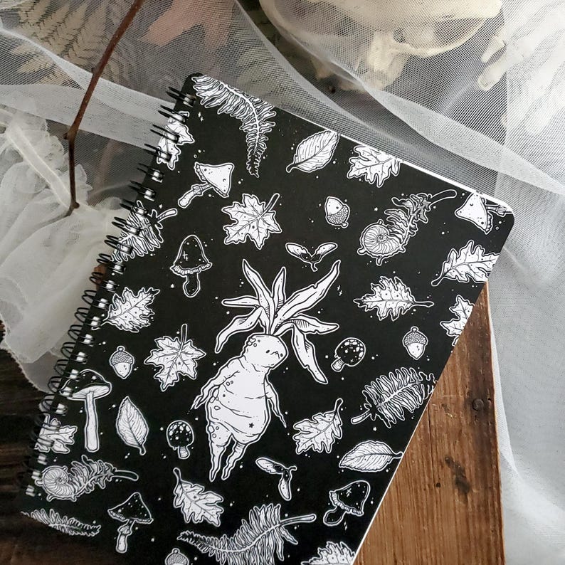 May include: A black spiral notebook featuring a white and black illustration of a woodland scene. The illustration includes mushrooms, leaves, acorns, and a creature with a human-like face and a plant growing from its head.