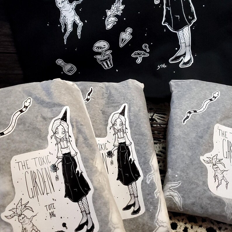 K&ouml;nnte beinhalten: Drei wei&szlig;e Stofftaschen mit Schwarz-Wei&szlig;-Illustrationen einer Hexe, einer Pflanze und einer Schlange. Die Taschen tragen den Text "The Toxic Garden" und "Tote Bag".