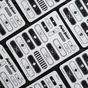 May include: A sheet of black and white band-aid stickers with various designs, including a skull, crossbones, heart, crescent moon, and stripes. The stickers are labeled "Bandages" and "Paper Haunt".