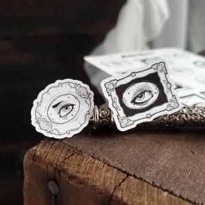 May include: Two black and white stickers with an eye design. The stickers are on a wooden surface. One sticker is a round frame with an eye looking to the left. The other sticker is a square frame with an eye looking to the right.