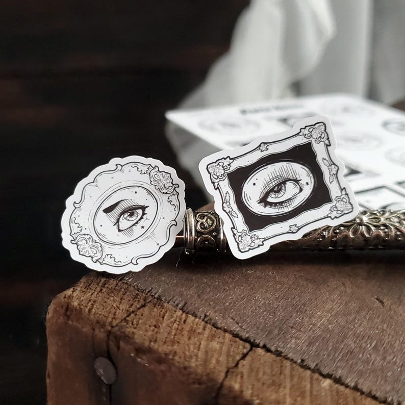 May include: Two black and white stickers with an eye design. The stickers are on a wooden surface. One sticker is a round frame with an eye looking to the left. The other sticker is a square frame with an eye looking to the right.