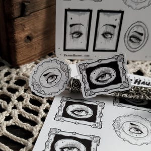 May include: A sheet of stickers featuring black and white illustrations of eyes in ornate frames. The stickers are printed on white paper and have a glossy finish. The text "PAPER HAUNT.COM" is printed at the bottom of the sheet.