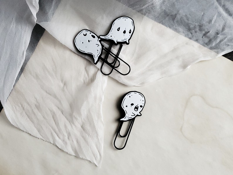 May include: Three black metal ghost-shaped paper clips with white ghost faces. The ghosts have different expressions. The paper clips are on a white surface.