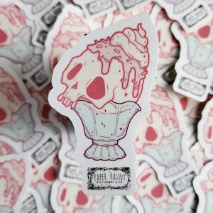 May include: A sticker depicting a skull-shaped sundae with pink and white hues, adorned with sprinkles and whipped cream. The sundae is presented in a decorative glass. The sticker includes the text "PAPER HAUNT STATIONERY & CO."