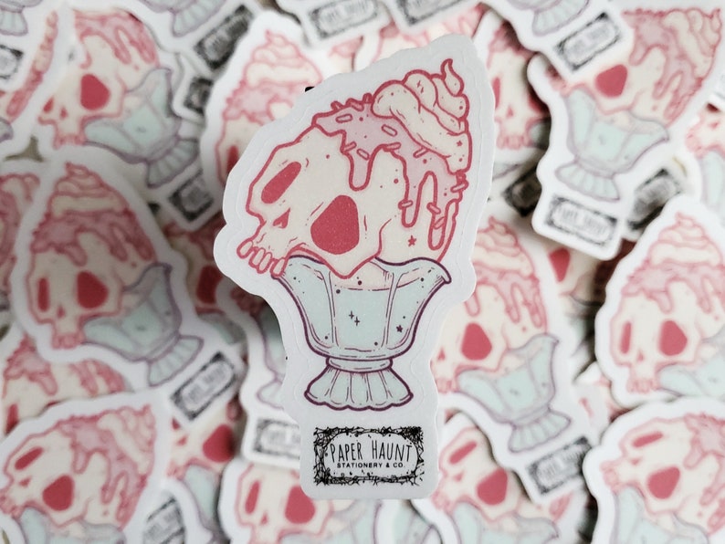 May include: A sticker depicting a skull-shaped sundae with pink and white hues, adorned with sprinkles and whipped cream. The sundae is presented in a decorative glass. The sticker includes the text "PAPER HAUNT STATIONERY & CO."
