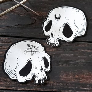 Op de afbeelding: Twee zwart-witte schedelpatches met geborduurde details. De ene schedel heeft een halve maan boven zijn oog en de andere heeft een pentagram op zijn voorhoofd.