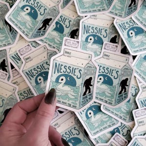 K&ouml;nnte beinhalten: Ein Stapel wei&szlig;er und blauer Aufkleber mit einer Cartoon-Illustration eines Nessie-&auml;hnlichen Wesens mit dem Text "Nessie's Missing" und "Have you seen me?" Die Aufkleber sind in Form eines Milchkartons gestaltet und haben einen blau-wei&szlig;en Hintergrund.