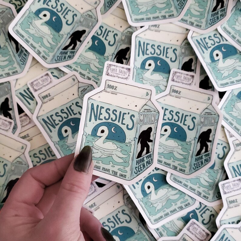 K&ouml;nnte beinhalten: Ein Stapel wei&szlig;er und blauer Aufkleber mit einer Cartoon-Illustration eines Nessie-&auml;hnlichen Wesens mit dem Text "Nessie's Missing" und "Have you seen me?" Die Aufkleber sind in Form eines Milchkartons gestaltet und haben einen blau-wei&szlig;en Hintergrund.
