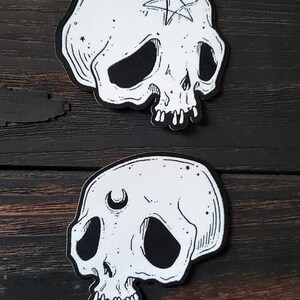 Op de afbeelding: Twee witte stoffen schedelpatches met zwarte lijntekeningen. De bovenste schedel heeft een vijfpuntige ster met een cirkel in het midden op zijn voorhoofd. De onderste schedel heeft een halve maan op zijn voorhoofd.