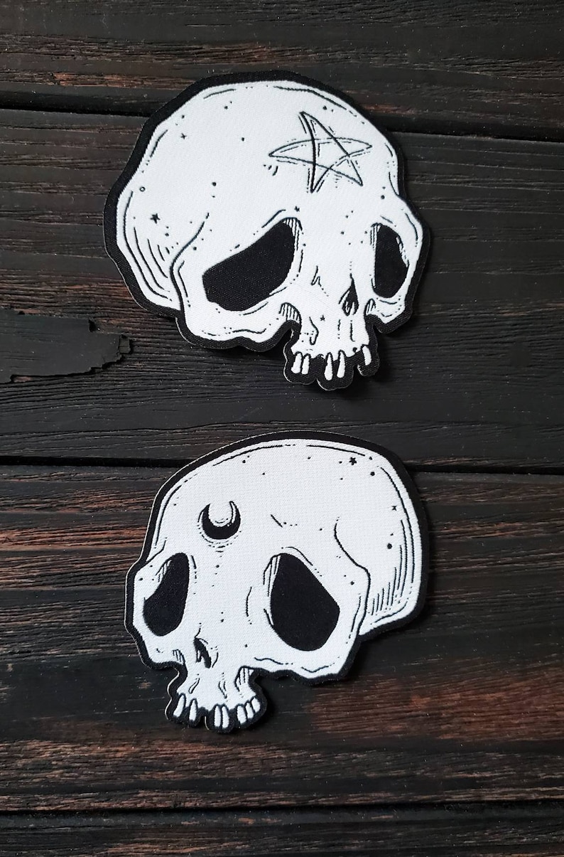 Op de afbeelding: Twee witte stoffen schedelpatches met zwarte lijntekeningen. De bovenste schedel heeft een vijfpuntige ster met een cirkel in het midden op zijn voorhoofd. De onderste schedel heeft een halve maan op zijn voorhoofd.