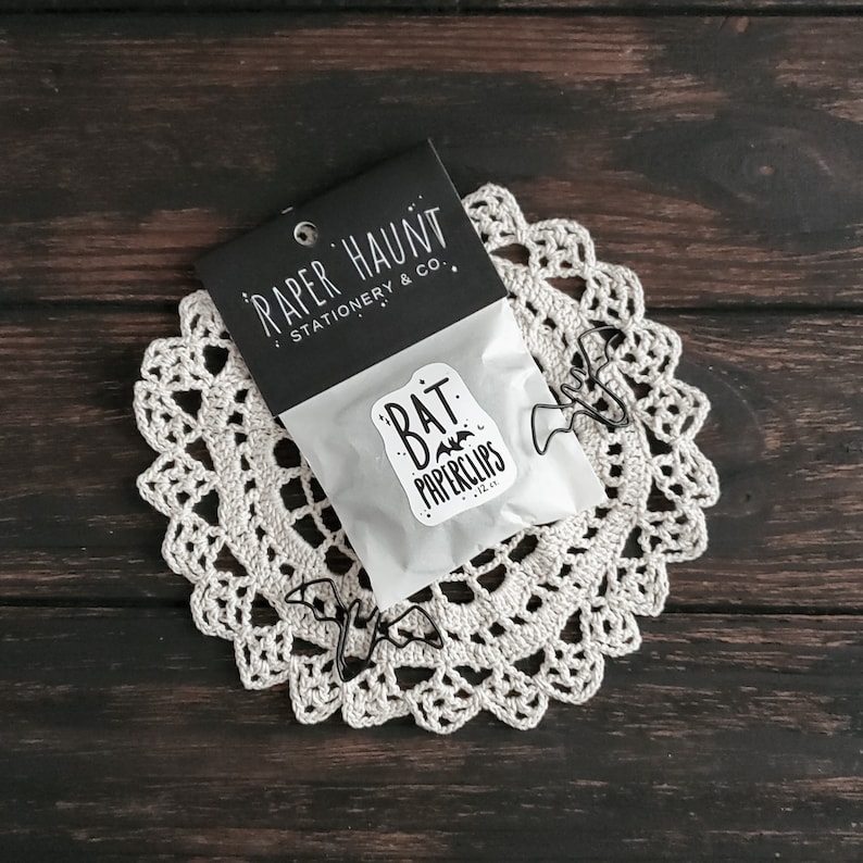 May include: A set of 12 black bat-shaped paperclips in a clear plastic bag with a white sticker that says "Bat Paperclips" on a white doily. The bag is on top of a black card that says "Paper Haunt Stationery & Co."