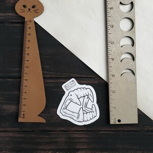 May include: A black and white illustration of a set of fangs on a white sticker. The sticker is placed on a wooden surface next to a brown cat-shaped ruler and a silver ruler with moon cutouts.