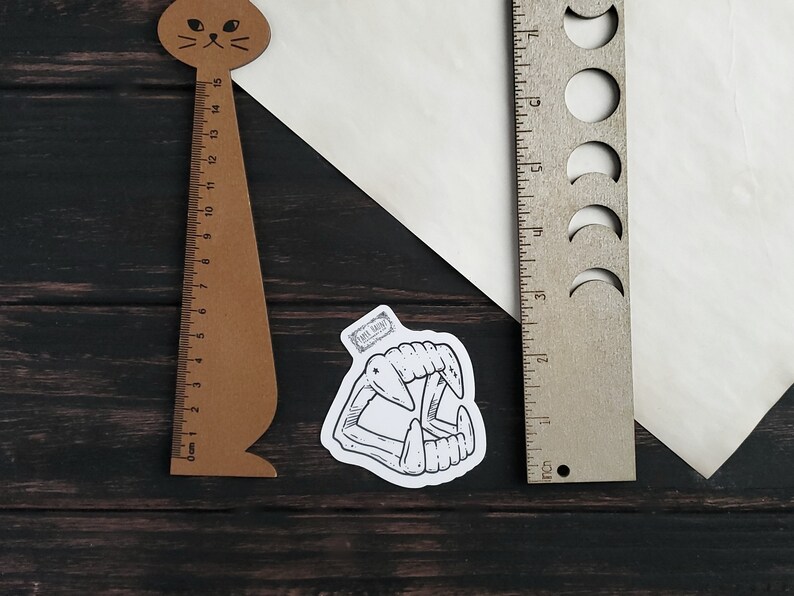 May include: A black and white illustration of a set of fangs on a white sticker. The sticker is placed on a wooden surface next to a brown cat-shaped ruler and a silver ruler with moon cutouts.