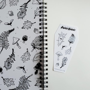 May include: A black and white sticker sheet with a coffin-shaped design. The sticker sheet features various illustrations of mushrooms, leaves, and other forest-themed elements. The text "PAPER HAUNT" is printed on the sticker sheet.