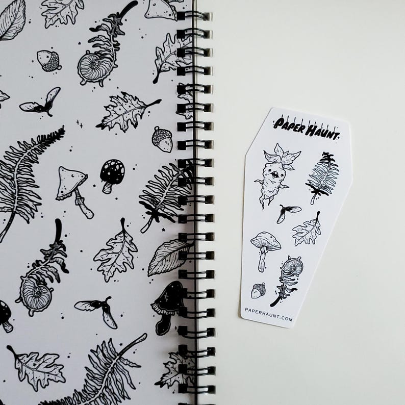 May include: A black and white sticker sheet with a coffin-shaped design. The sticker sheet features various illustrations of mushrooms, leaves, and other forest-themed elements. The text "PAPER HAUNT" is printed on the sticker sheet.