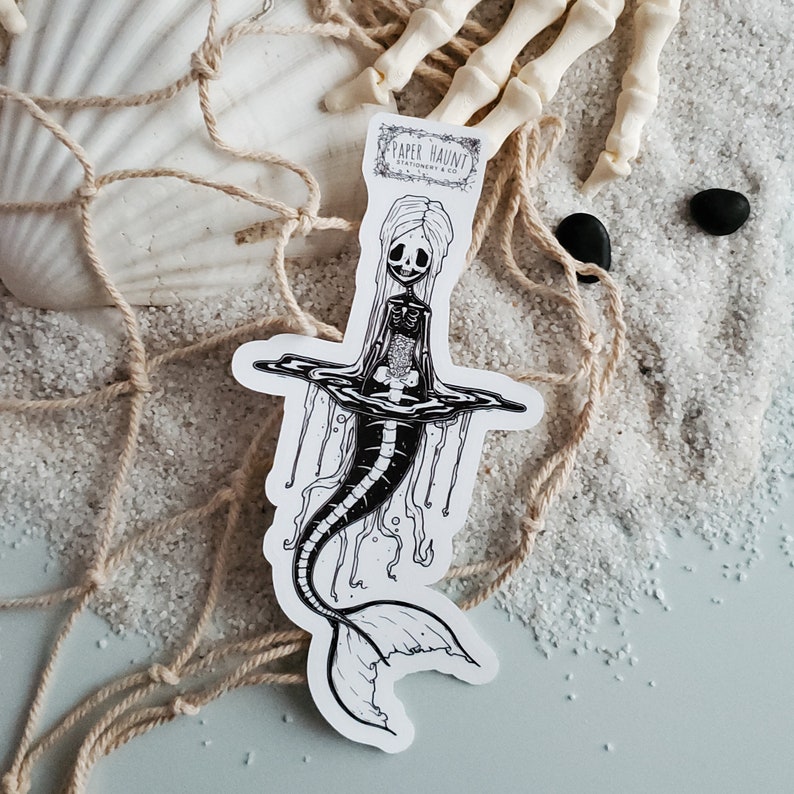 Spooky cute goth Jelly bones mermaid skeleton emerging from water, a sticker Featuring a Mermaid with Translucent skin with visible skeleton and organs with a sea shell on sand.