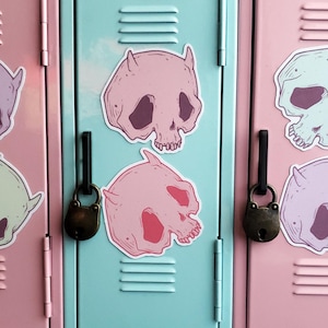 May include: Pink skull stickers with horns are affixed to the front of a light blue locker. A second pink skull sticker is below the first. A brass padlock hangs from the locker. Additional stickers are visible on adjacent pink lockers.
