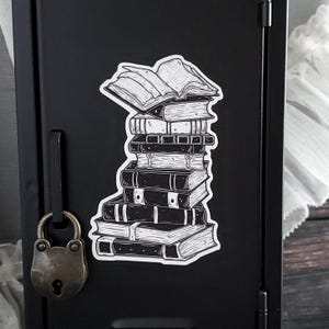 May include: Black and white sticker featuring a stack of books. The books are of varying sizes and styles, with one open at the top. The sticker is placed on a black surface, suggesting a theme of literature or reading.