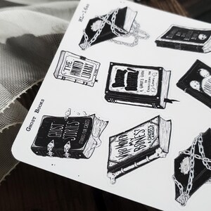 May include: A set of nine black and white stickers featuring illustrations of books with spooky themes. The titles include "The Haunted Library", "Ghost Stories", "A Ghost's Guide to Exorcising the Living", "Where  My Bones?", and "A Ghost's Lament".