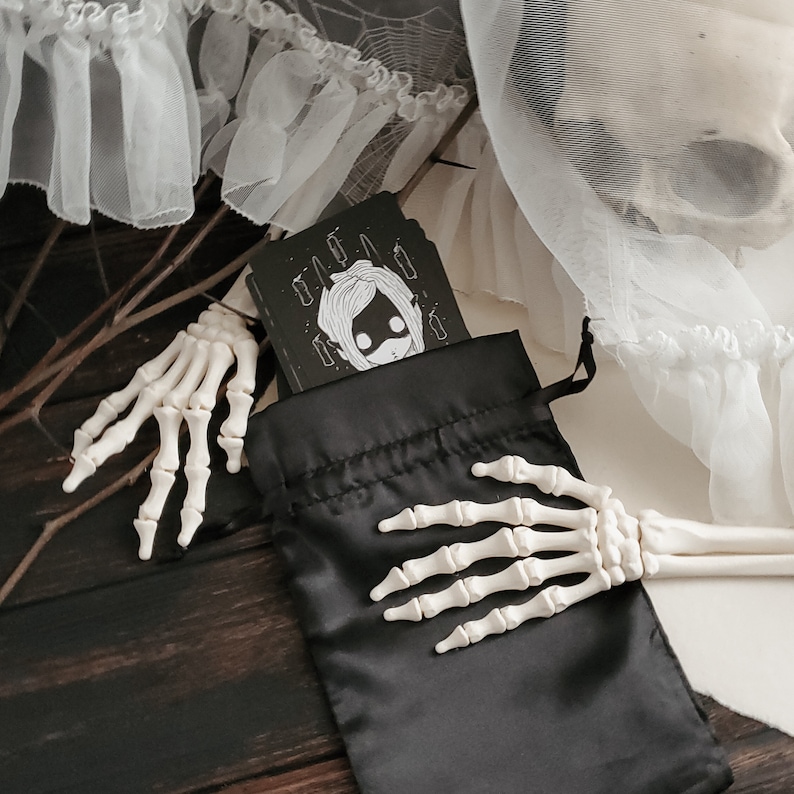 May include: A black drawstring bag with a white and black illustration of a girl with a crown on her head. The bag is held by a skeletal hand.