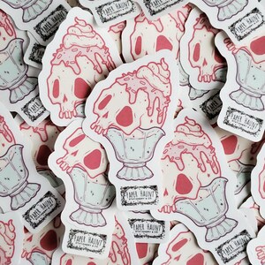 May include: A pile of stickers depicting a skull-shaped ice cream sundae with pink ice cream and sprinkles. The stickers are in a light pink, white, and mint green colour scheme. The text "PAPER HAUNT STATIONERY & CO." is printed on a black label.