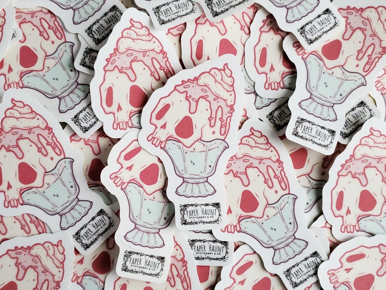 May include: A pile of stickers depicting a skull-shaped ice cream sundae with pink ice cream and sprinkles. The stickers are in a light pink, white, and mint green colour scheme. The text "PAPER HAUNT STATIONERY & CO." is printed on a black label.