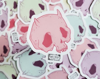 May include: A pink skull sticker with red horns and details. The sticker is surrounded by other stickers in various pastel colors. The sticker has the text "PAPER HAUNT STATIONERY & CO." printed on the bottom.