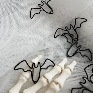 May include: Black metal bat paper clips on a white tulle background. The paper clips are shaped like bats and are arranged in a random pattern.
