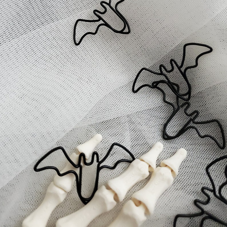 May include: Black metal bat paper clips on a white tulle background. The paper clips are shaped like bats and are arranged in a random pattern.
