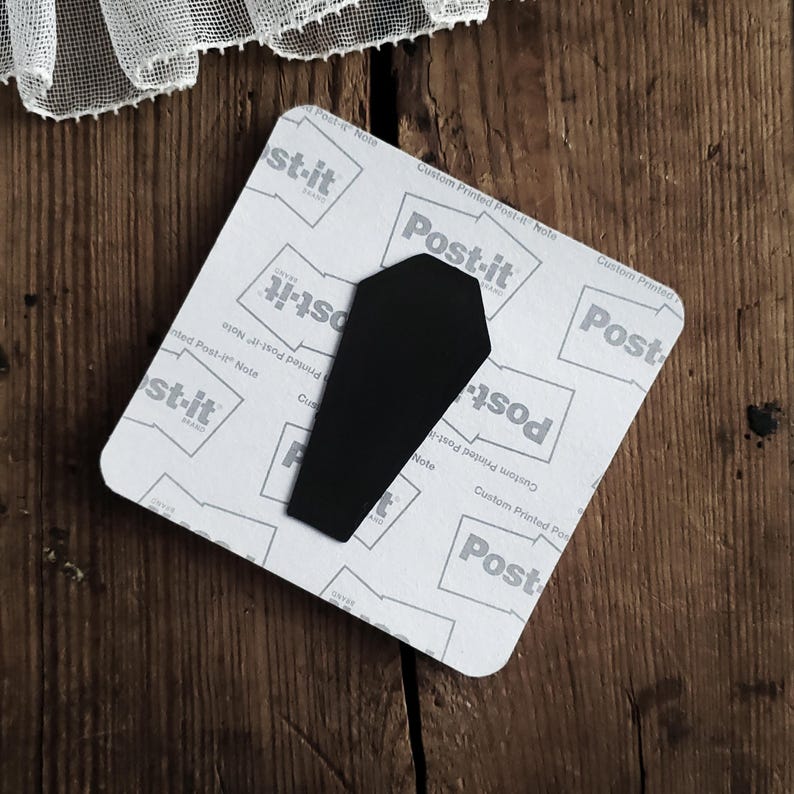 May include: A black coffin-shaped magnet is attached to a Post-it Note. The Post-it Note is white with gray text and a square shape. The background is a dark wood surface.
